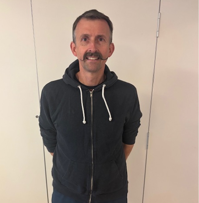 En mann med kort hår og bart står mot en ensfarget, lys vegg, iført en mørk hettegenser med glidelås og hvite snorer.