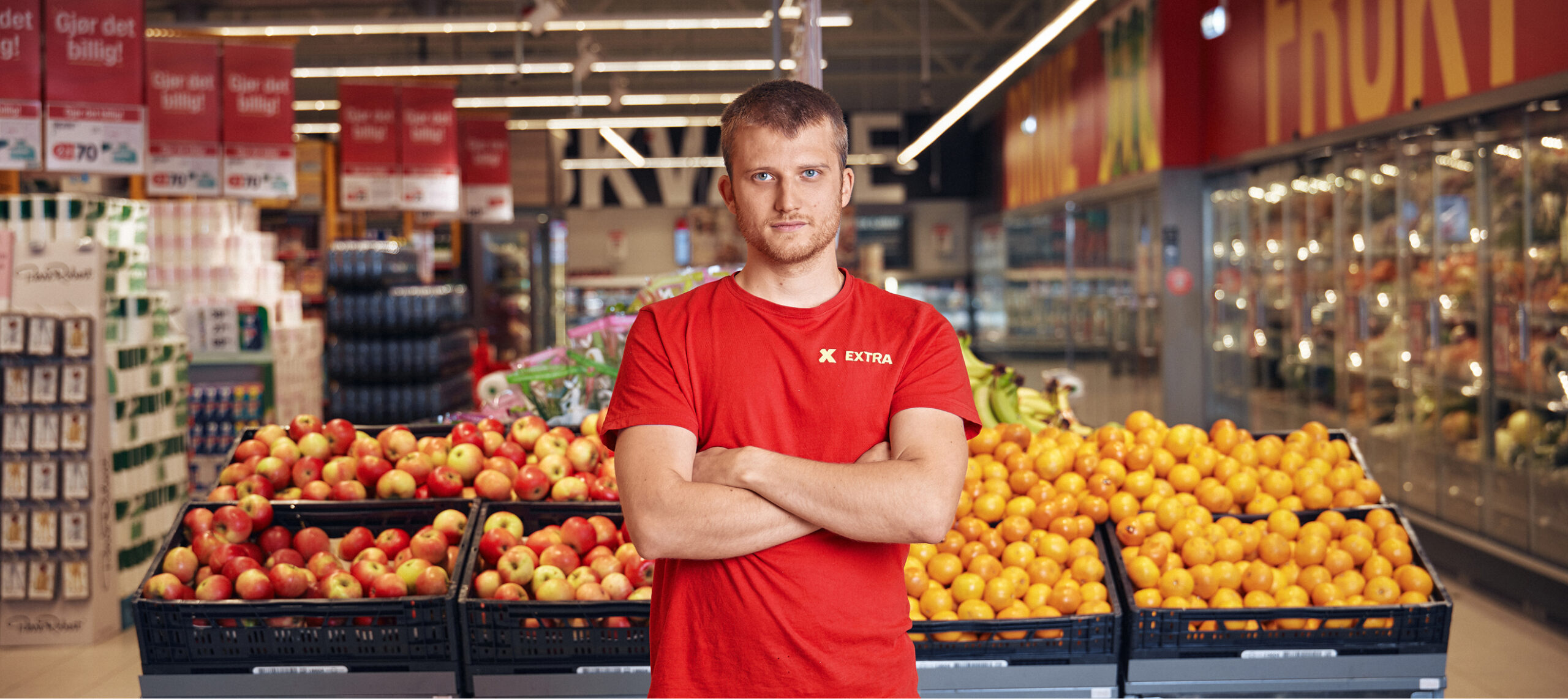 En mann i rød skjorte står med armene i kors foran eple- og appelsinutstillinger inne i en dagligvarebutikk.